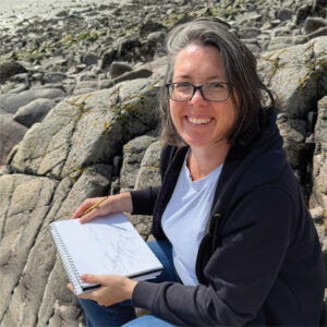 Woman sitting on rocks by the beach, sketching in a notebook and smiling at the camera.
