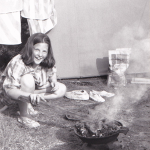 Old black and white photo of a young girl cooking outside on a BBQ.