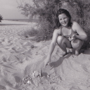 Old black and white photo of a young girl on a beach pointing at a collection of stones and a feather.