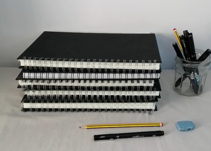 Image of a pile of sketch books, with a pen, pencil and rubber in front and more pens and pencils in a jam jar to the right.