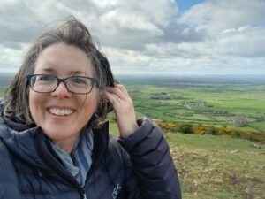 Woman smiling holding her hair away from her face, on the top of a hill with the countryside spreading out behind in the distance.