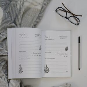 Image of an open Gratitude Journal beside a grey blanket, pen and reading glasses.