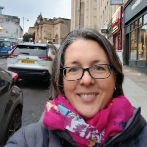 woman with greying hair and glasses, standing on a city street smiling at the camera.