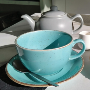Teal cup, saucer and pale grey teapot on a white table in bright sunlight.