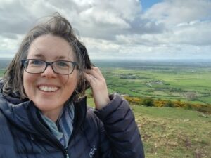 Woman smiling holding her hair away from her face, on the top of a hill with the countryside spreading out behind in the distance.