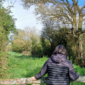 Photo of the back of a woman standing next to a wooden gate looking into the UK Spring countryside beyond.